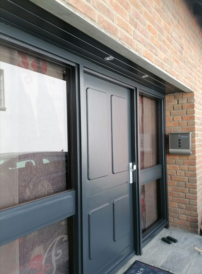 Eures • Moderne Eingangstür in dunkler Farbe, umgeben von Ziegelmauer.